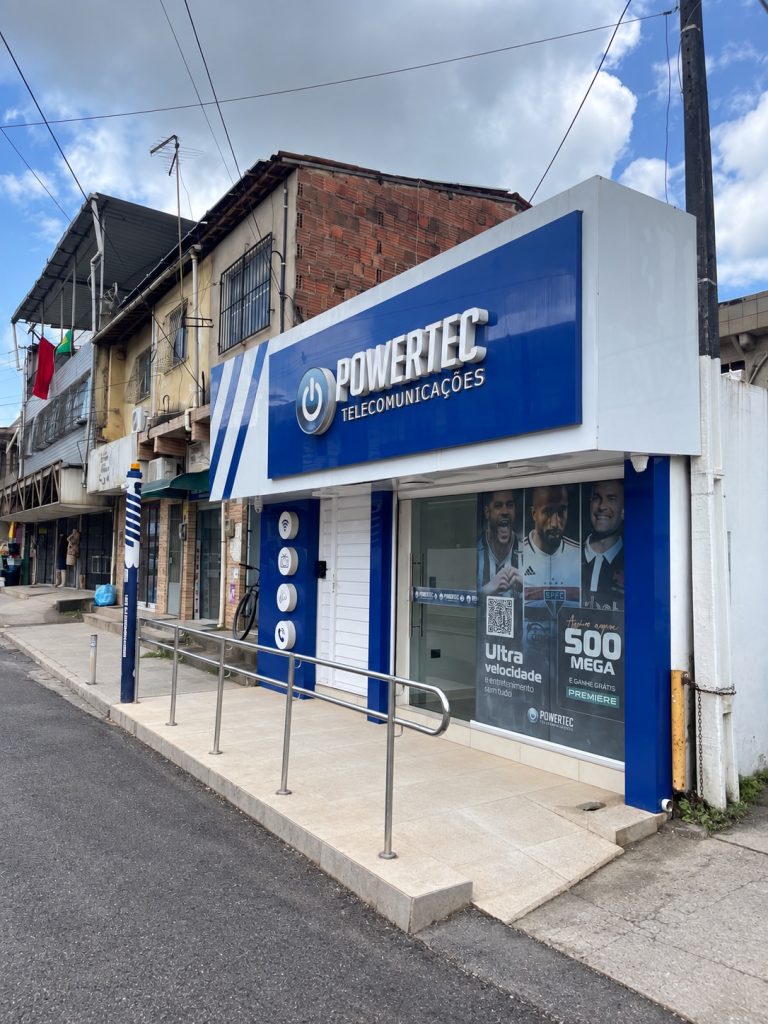 Vista lateral da loja Powertec Telecomunicações, com fachada azul e branca e rampa de acesso.